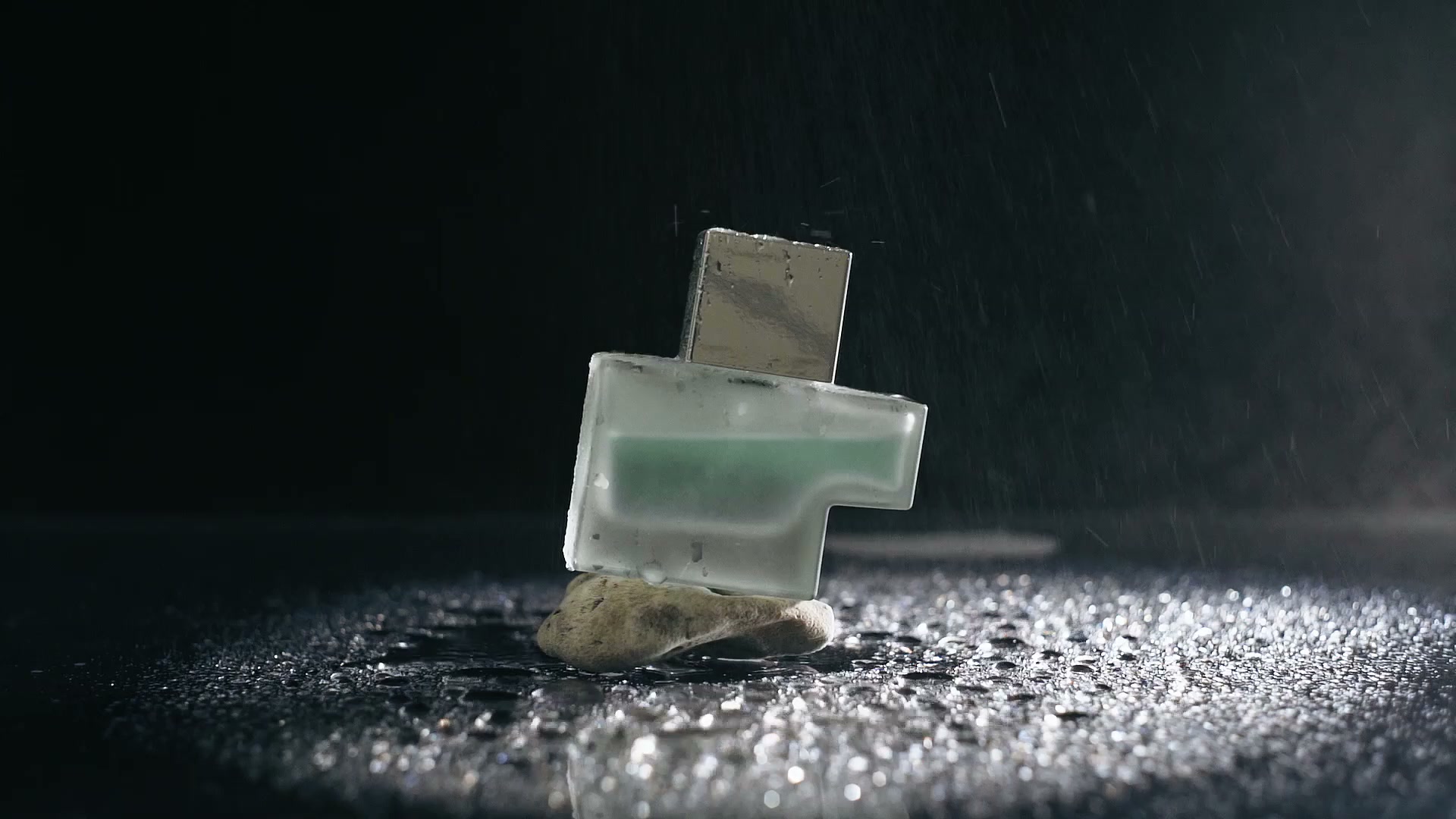 A frosted perfume bottle rests on wet stone under falling water on a dark surface.
